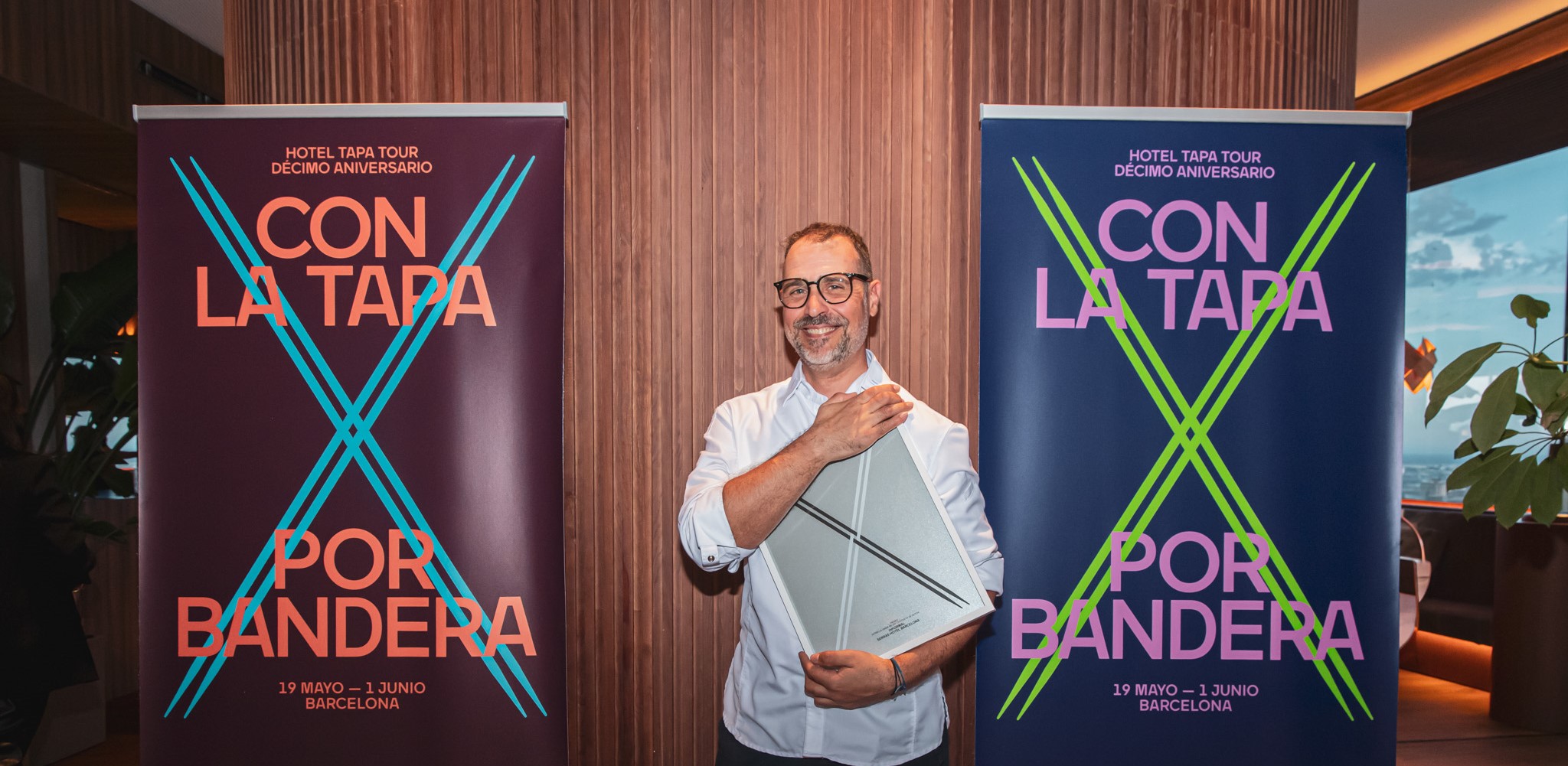 Chef sosteniendo el certificado del premio, de pie frente a dos coloridos roll-ups del evento Hotel Tapa Tour